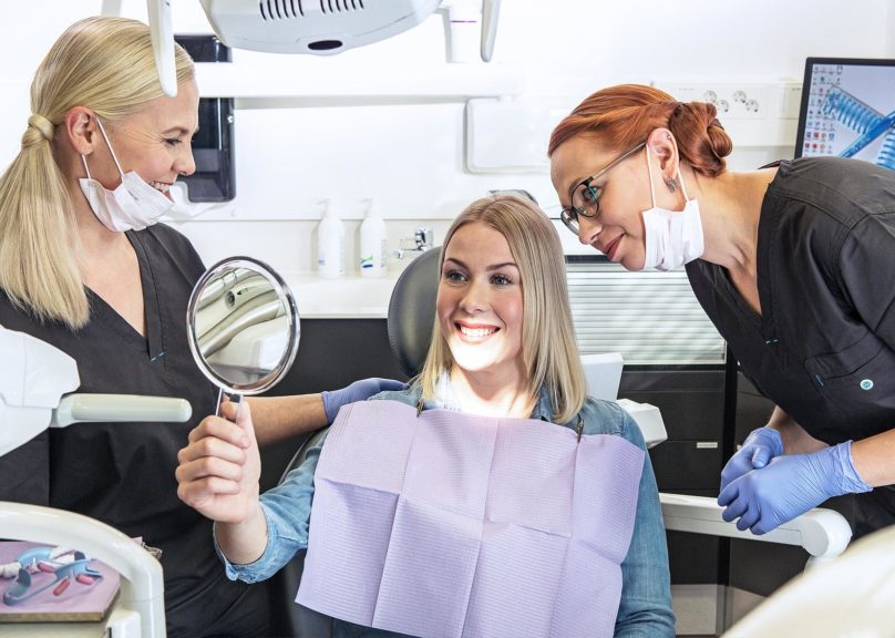 Työterveyshammashuollon naispuolinen asiakas katsoo ihaillen peiliin vierellään Coronarian hammaslääkäri ja hammashoitaja.
